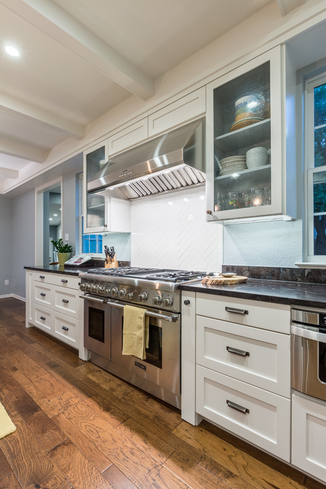 Example of a mid-sized transitional l-shaped medium tone wood floor eat-in kitchen design in Austin with an undermount sink, shaker cabinets, white cabinets, granite countertops, white backsplash, ceramic backsplash, stainless steel appliances and an island