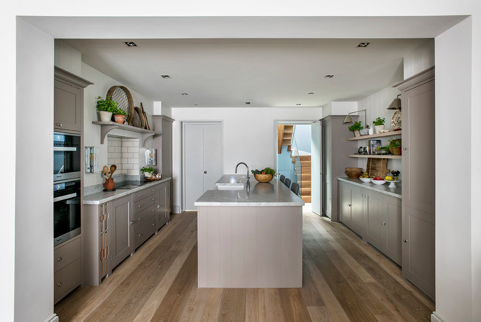 Inspiration for a large traditional galley separate kitchen with a farmhouse sink, shaker cabinets, brown cabinets, marble benchtops, beige splashback, subway tile splashback, stainless steel appliances, medium hardwood floors, with island, brown floor and grey benchtop.