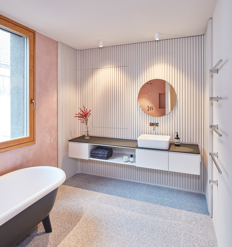 This is an example of a large contemporary bathroom in Munich with louvered cabinets, white cabinets, a freestanding tub, a curbless shower, a wall-mount toilet, pink walls, terrazzo floors, a vessel sink, wood benchtops, grey floor, an open shower and black benchtops.