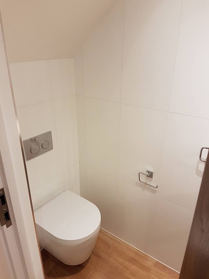 This is an example of a mid-sized modern kids bathroom in London with a wall-mount toilet, white tile, ceramic tile, white walls, laminate floors and brown floor.