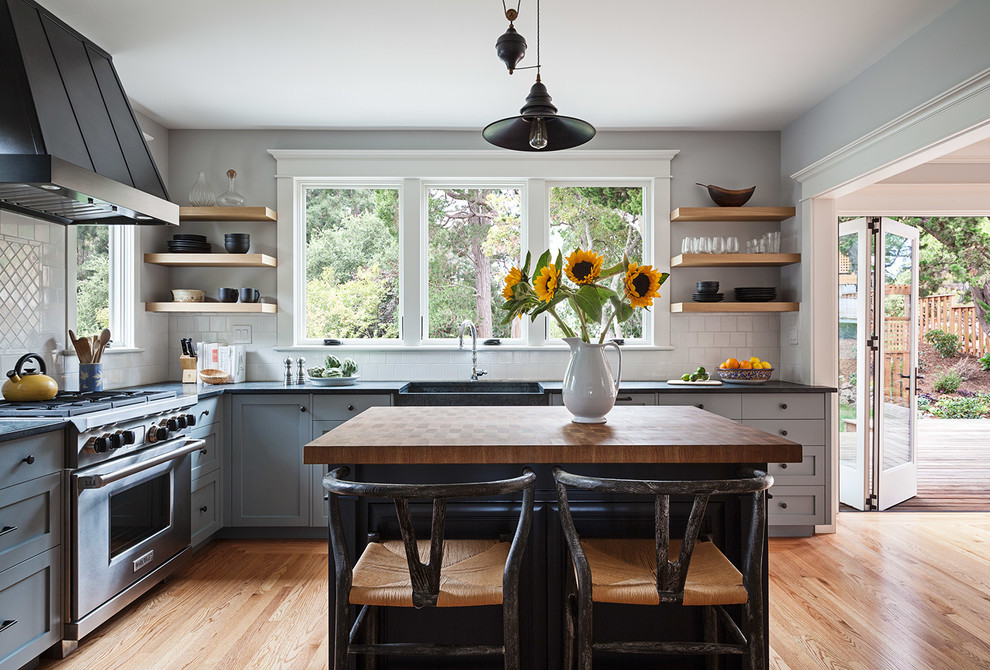 This is an example of a mid-sized arts and crafts l-shaped kitchen in San Francisco with a farmhouse sink, shaker cabinets, grey cabinets, white splashback, ceramic splashback, stainless steel appliances, light hardwood floors, with island, beige floor and soapstone benchtops.