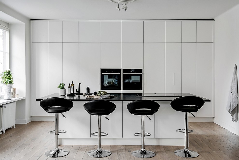 Photo of a scandinavian kitchen in Melbourne with flat-panel cabinets, white cabinets, black appliances, light hardwood floors, with island, beige floor and black benchtop.