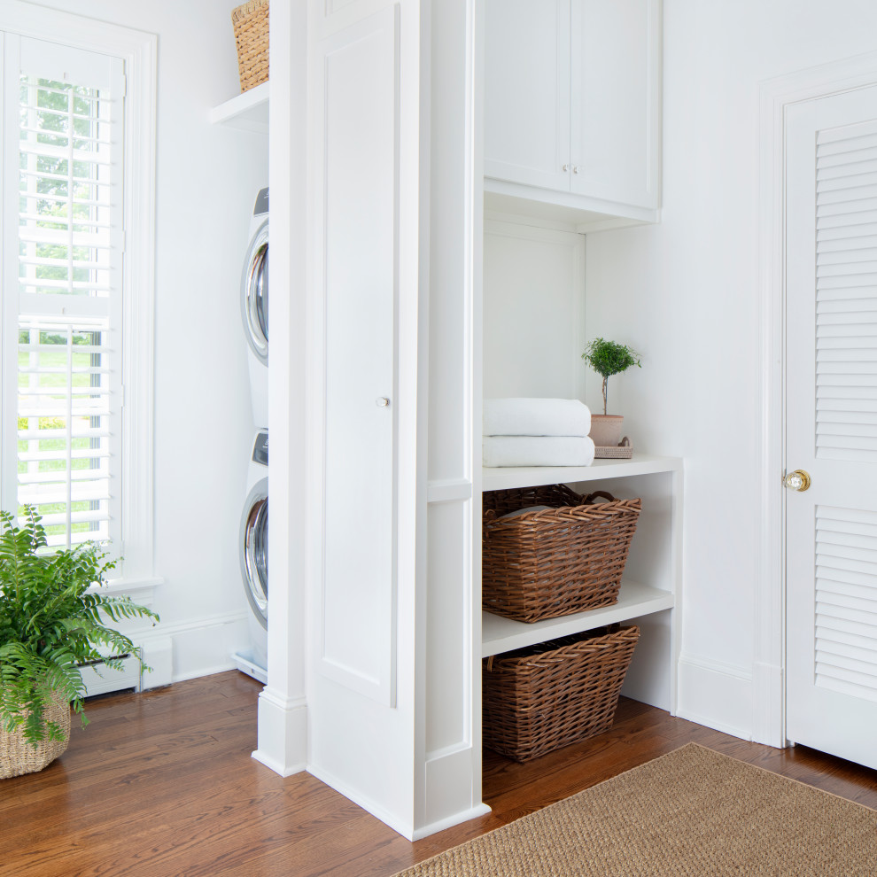 Mid-sized traditional laundry cupboard in Richmond with flat-panel cabinets, white cabinets, white walls, medium hardwood floors, a stacked washer and dryer and brown floor.