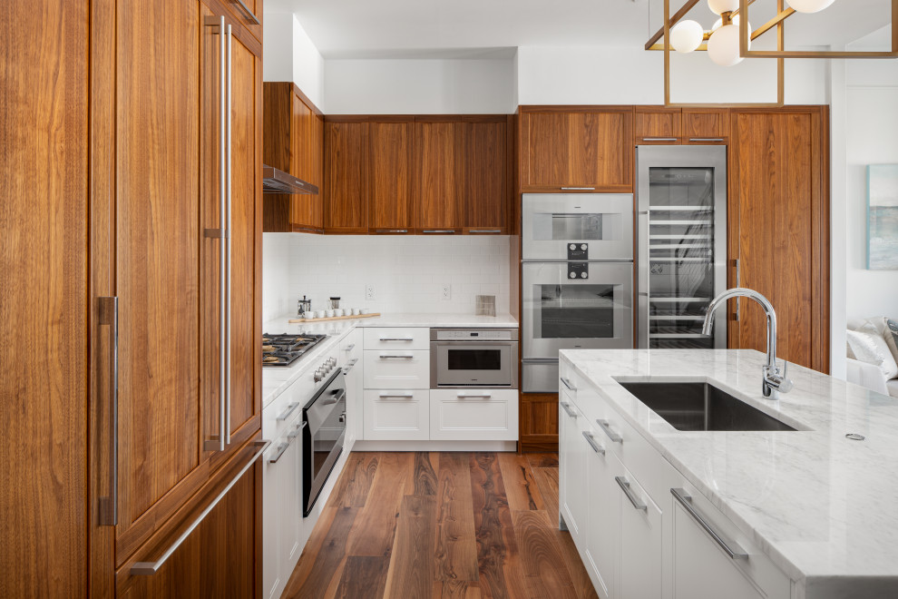 Large contemporary l-shaped open plan kitchen in Providence with an undermount sink, shaker cabinets, medium wood cabinets, white splashback, subway tile splashback, stainless steel appliances, medium hardwood floors, with island, brown floor and white benchtop.