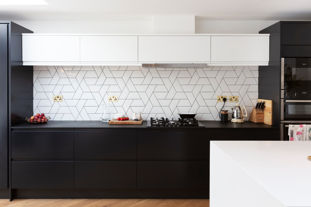 Design ideas for a large contemporary galley open plan kitchen in London with flat-panel cabinets, solid surface benchtops, white splashback, ceramic splashback, black appliances, medium hardwood floors, with island, black benchtop and brown floor.