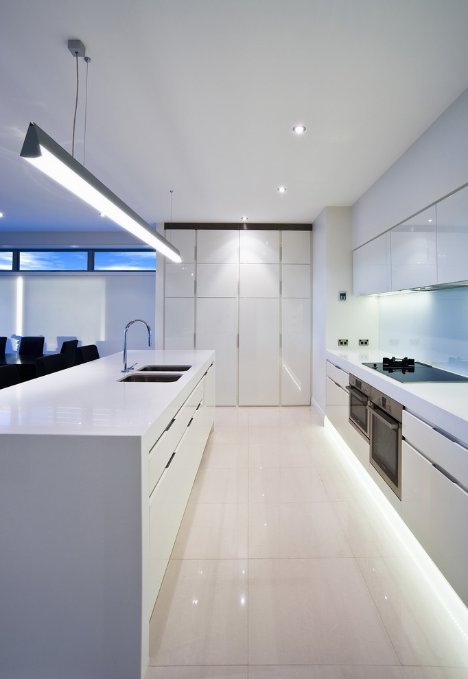 This is an example of a modern galley open plan kitchen in Auckland with an undermount sink, flat-panel cabinets, white cabinets, quartz benchtops, white splashback, glass sheet splashback, stainless steel appliances and white floor.