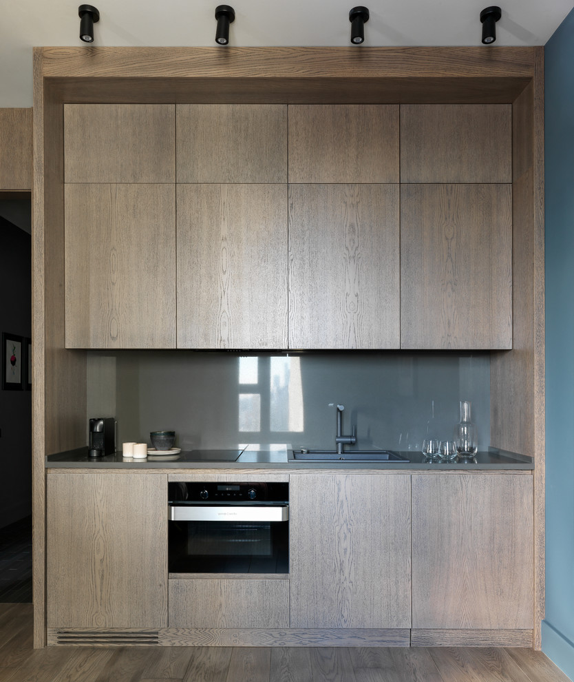 Photo of a small contemporary single-wall open plan kitchen in Moscow with flat-panel cabinets, medium wood cabinets, quartz benchtops, grey splashback, black appliances, medium hardwood floors, no island, brown floor, grey benchtop and a drop-in sink.