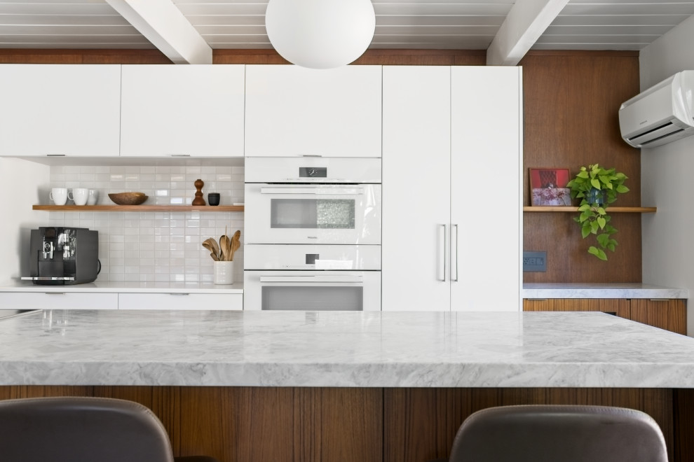 This is an example of a large midcentury l-shaped open plan kitchen in Orange County with an undermount sink, flat-panel cabinets, white cabinets, quartzite benchtops, white splashback, ceramic splashback, white appliances, slate floors, with island, grey floor, multi-coloured benchtop and timber.