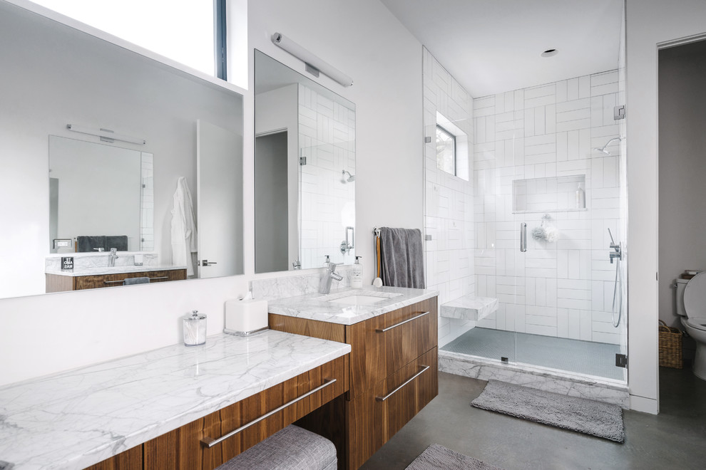 Design ideas for a mid-sized contemporary master bathroom in Austin with flat-panel cabinets, an alcove shower, a two-piece toilet, white tile, white walls, concrete floors, an undermount sink, a hinged shower door, medium wood cabinets, stone tile, marble benchtops, grey floor, an alcove tub and grey benchtops.