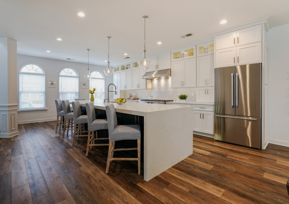 Photo of a large classic single-wall kitchen/diner in DC Metro with a belfast sink, recessed-panel cabinets, white cabinets, engineered stone countertops, white splashback, glass tiled splashback, black appliances, dark hardwood flooring, an island and white worktops.