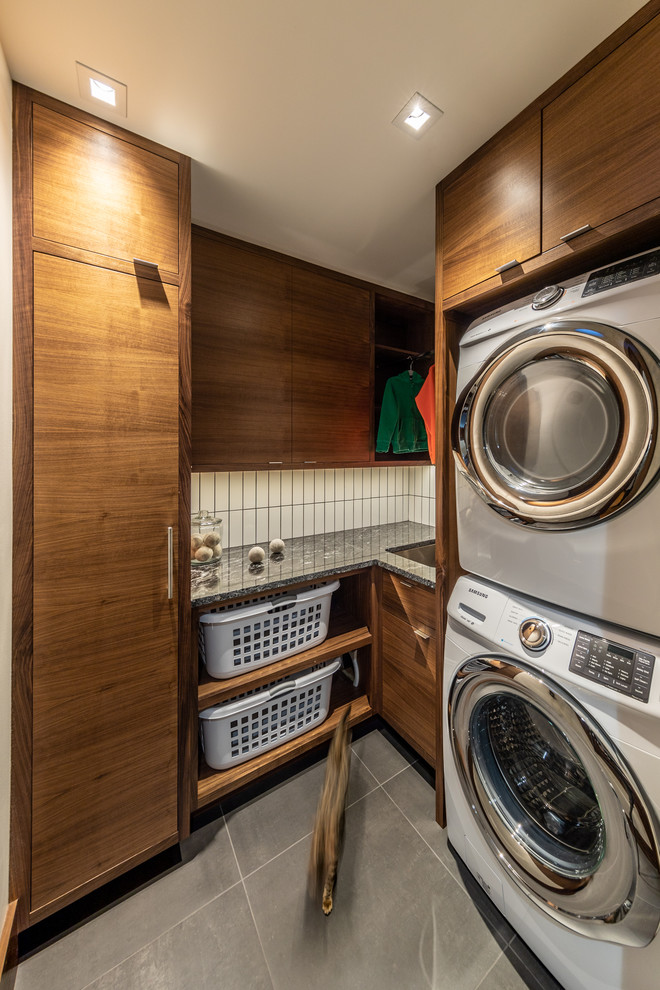 This is an example of a small modern l-shaped dedicated laundry room in Cedar Rapids with a drop-in sink, flat-panel cabinets, dark wood cabinets, granite benchtops, white walls, porcelain floors, a stacked washer and dryer, grey floor and black benchtop.