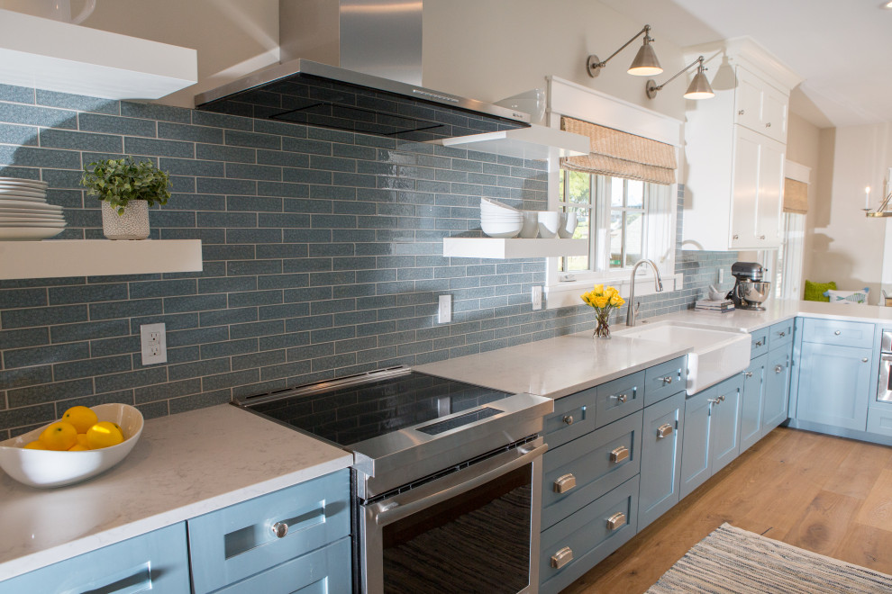 This is an example of a mid-sized beach style l-shaped eat-in kitchen in Other with a farmhouse sink, flat-panel cabinets, blue cabinets, quartz benchtops, blue splashback, glass tile splashback, panelled appliances, light hardwood floors, no island, beige floor and white benchtop.