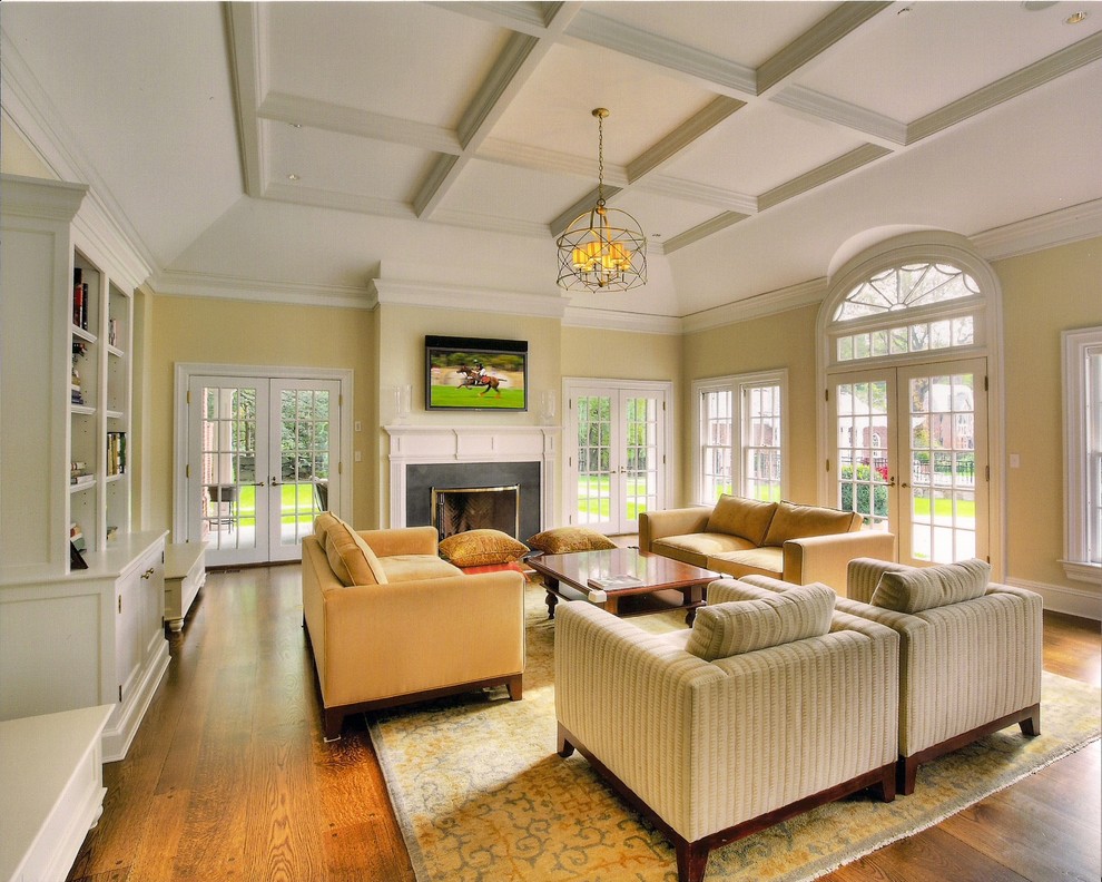 This is an example of a large traditional family room in New York with a library, yellow walls, medium hardwood floors, a standard fireplace, a wood fireplace surround and a wall-mounted tv.