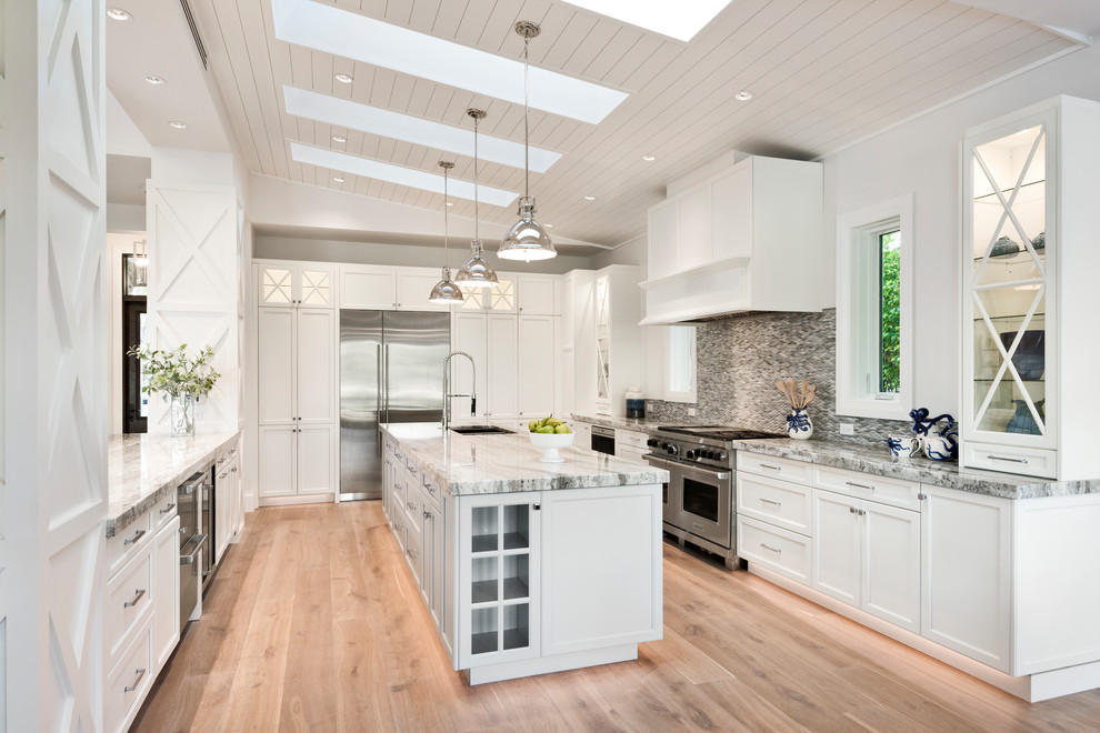Large beach style u-shaped open plan kitchen in Other with recessed-panel cabinets, white cabinets, granite benchtops, grey splashback, stainless steel appliances, medium hardwood floors, brown floor, an undermount sink and with island.