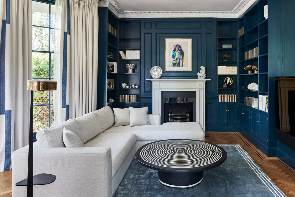 Photo of a mid-sized transitional enclosed family room in London with a library, blue walls, a standard fireplace, a stone fireplace surround, medium hardwood floors, a wall-mounted tv and brown floor.