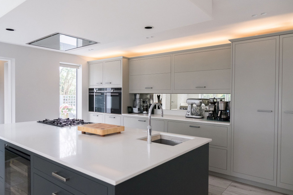Mid-sized contemporary single-wall open plan kitchen in Hertfordshire with a drop-in sink, flat-panel cabinets, grey cabinets, solid surface benchtops, metallic splashback, mirror splashback, panelled appliances, porcelain floors, with island, grey floor and white benchtop.