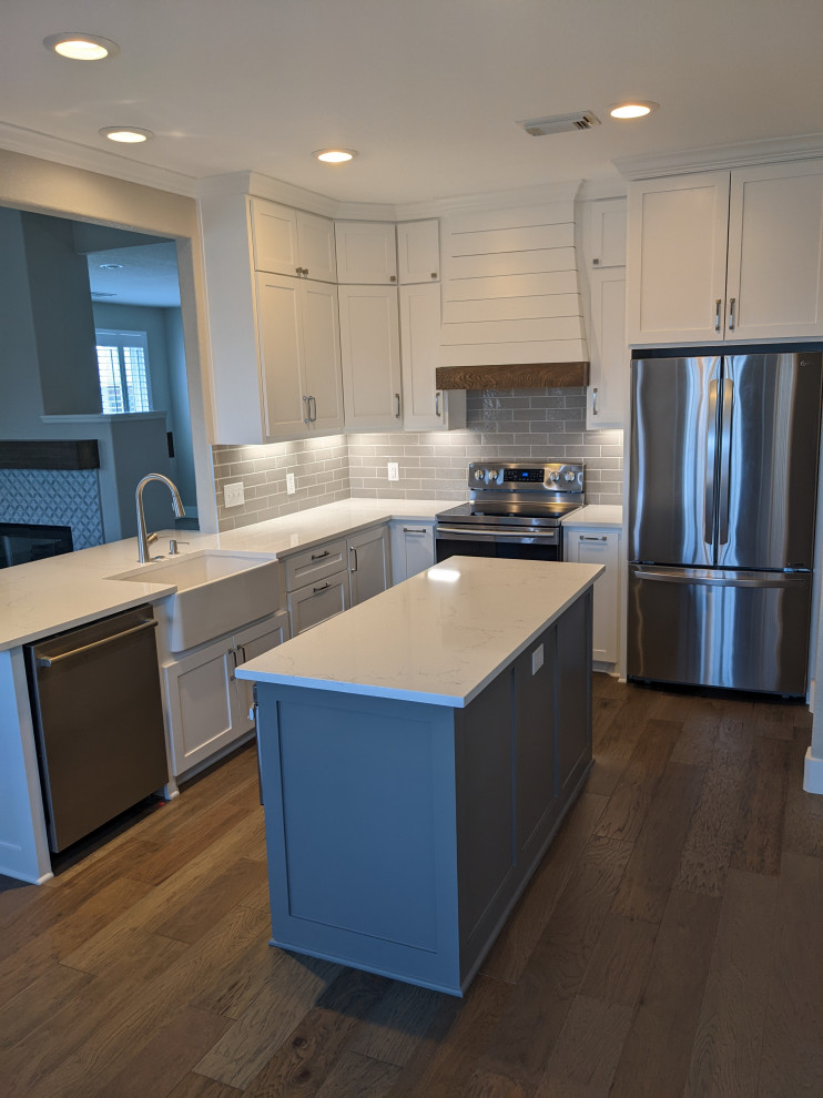 Mid-sized country u-shaped kitchen in Houston with a farmhouse sink, shaker cabinets, white cabinets, quartz benchtops, grey splashback, subway tile splashback, stainless steel appliances, medium hardwood floors, with island, brown floor and white benchtop.