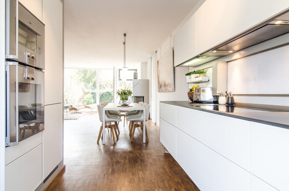This is an example of a mid-sized scandinavian galley open plan kitchen in Frankfurt with flat-panel cabinets, white cabinets, white splashback, stainless steel appliances, bamboo floors, no island, brown floor and black benchtop.