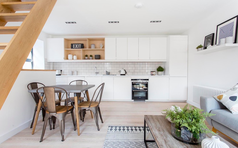 This is an example of a mid-sized scandinavian single-wall open plan kitchen in Barcelona with light hardwood floors, flat-panel cabinets, white cabinets, white splashback, subway tile splashback, black appliances and no island.