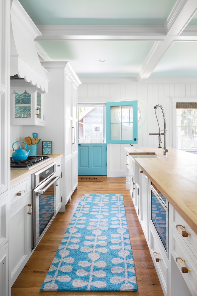 Inspiration for a beach style galley open plan kitchen in Boston with a farmhouse sink, recessed-panel cabinets, white cabinets, wood benchtops, white splashback, timber splashback, panelled appliances, medium hardwood floors and with island.