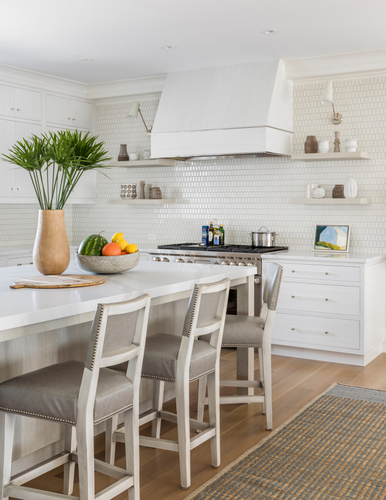 Beach style l-shaped kitchen in New York with white cabinets, white splashback, stainless steel appliances, light hardwood floors, with island and white benchtop.