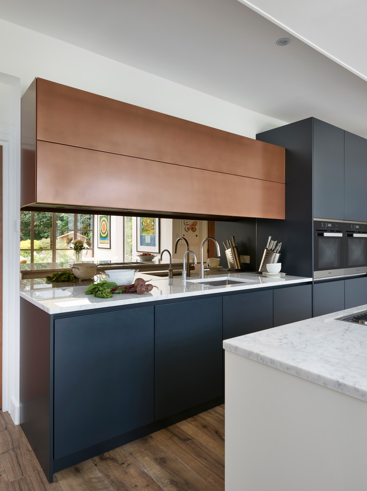 Large contemporary l-shaped eat-in kitchen in London with an integrated sink, blue cabinets, marble benchtops, metallic splashback, mirror splashback, light hardwood floors, with island, brown floor and white benchtop.
