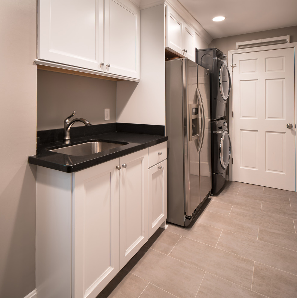 This is an example of a mid-sized transitional single-wall utility room in Charlotte with a single-bowl sink, white cabinets, quartz benchtops, grey walls, porcelain floors, a stacked washer and dryer and recessed-panel cabinets.