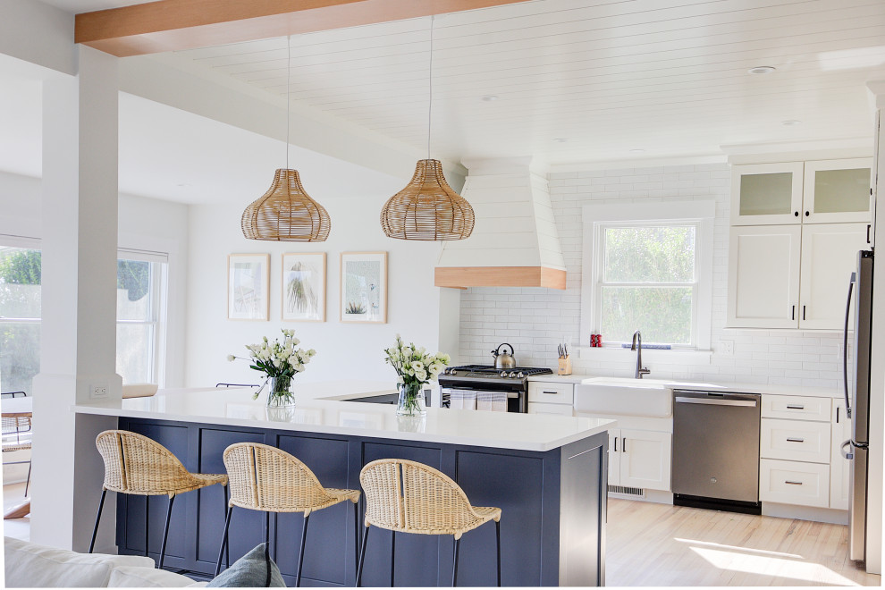 Inspiration for a mid-sized beach style u-shaped open plan kitchen in New York with a farmhouse sink, quartz benchtops, white splashback, ceramic splashback, stainless steel appliances, light hardwood floors, white benchtop, timber, shaker cabinets, white cabinets, a peninsula and beige floor.