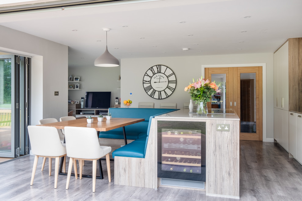 This is an example of a large scandinavian eat-in kitchen in Gloucestershire with a farmhouse sink, flat-panel cabinets, white cabinets, solid surface benchtops, blue splashback, glass sheet splashback, black appliances, porcelain floors, with island, brown floor and white benchtop.