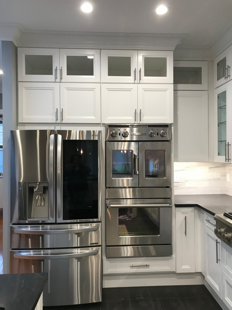 Large contemporary u-shaped kitchen pantry in New York with an undermount sink, recessed-panel cabinets, white cabinets, solid surface benchtops, grey splashback, stone tile splashback, stainless steel appliances, ceramic floors, with island, black floor, black benchtop and vaulted.