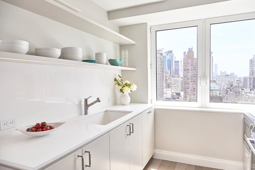 Photo of a small transitional u-shaped separate kitchen in New York with an undermount sink, flat-panel cabinets, white cabinets, solid surface benchtops, white splashback, stone slab splashback, stainless steel appliances, light hardwood floors, no island and beige floor.