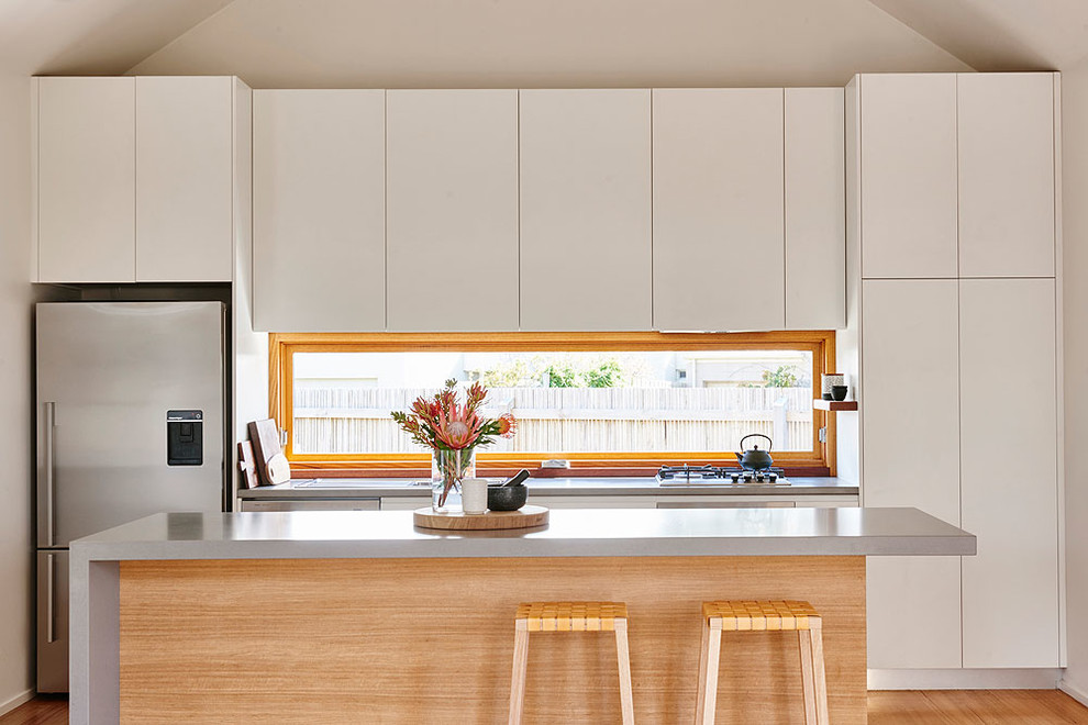 Photo of a beach style kitchen in Geelong with flat-panel cabinets, white cabinets, window splashback, stainless steel appliances, light hardwood floors, with island, beige floor and grey benchtop.