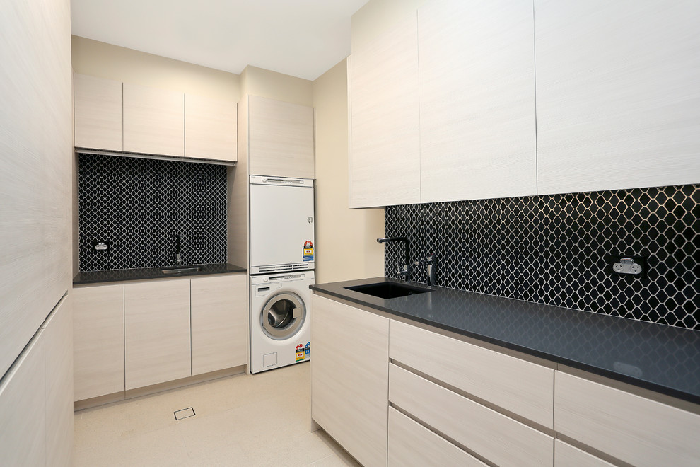 This is an example of a mid-sized galley utility room in Sydney with flat-panel cabinets, black splashback, stone slab splashback, beige cabinets, an undermount sink, quartz benchtops, beige walls, porcelain floors, a stacked washer and dryer and beige floor.