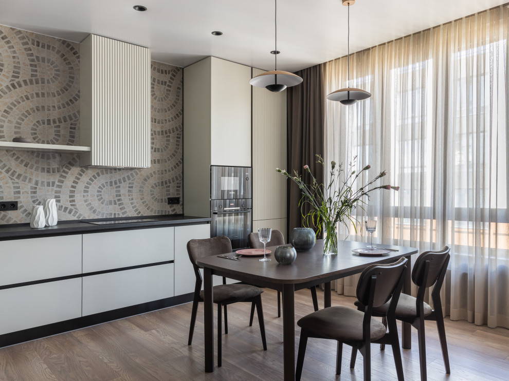 Foto på ett funkis grå linjärt kök och matrum, med släta luckor, vita skåp, flerfärgad stänkskydd, svarta vitvaror, mellanmörkt trägolv och brunt golv