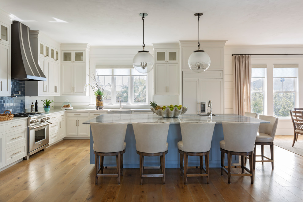 Photo of a beach style l-shaped kitchen in Charleston with a farmhouse sink, shaker cabinets, white cabinets, blue splashback, subway tile splashback, stainless steel appliances, medium hardwood floors, with island and grey benchtop.