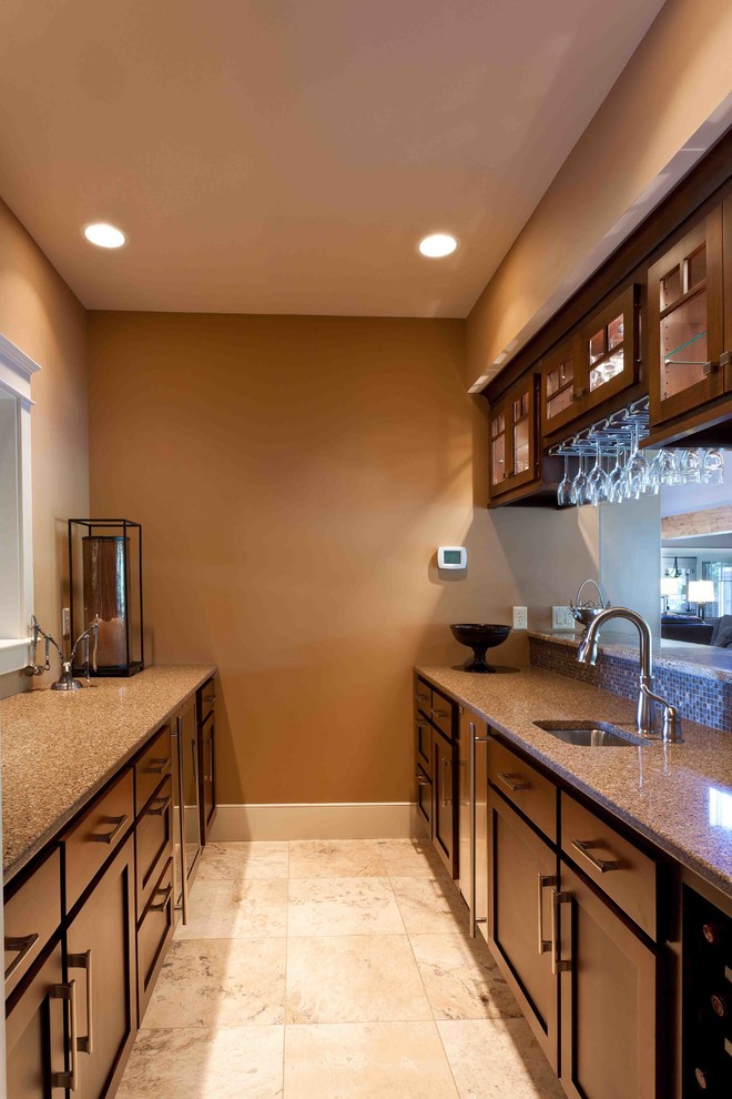 Photo of a mid-sized country galley wet bar in Other with recessed-panel cabinets, brown cabinets, granite benchtops, ceramic floors, beige floor and an undermount sink.