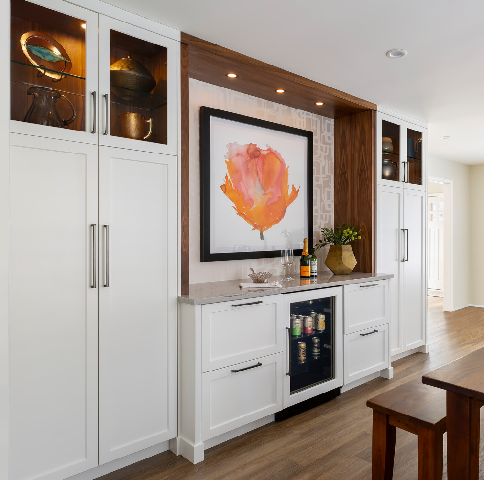 This is an example of a mid-sized transitional l-shaped home bar in Minneapolis with flat-panel cabinets, white cabinets, quartzite benchtops, grey splashback, stone slab splashback, vinyl floors, brown floor and grey benchtop.