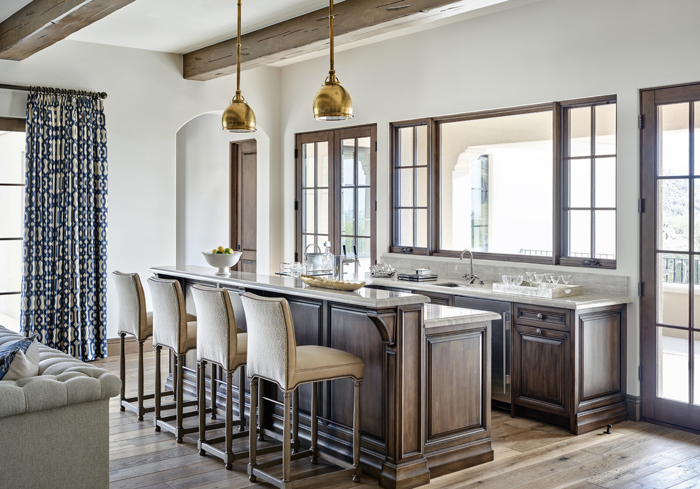 This is an example of a large mediterranean galley seated home bar in Chicago with an undermount sink, raised-panel cabinets, dark wood cabinets, light hardwood floors, marble benchtops and brown floor.