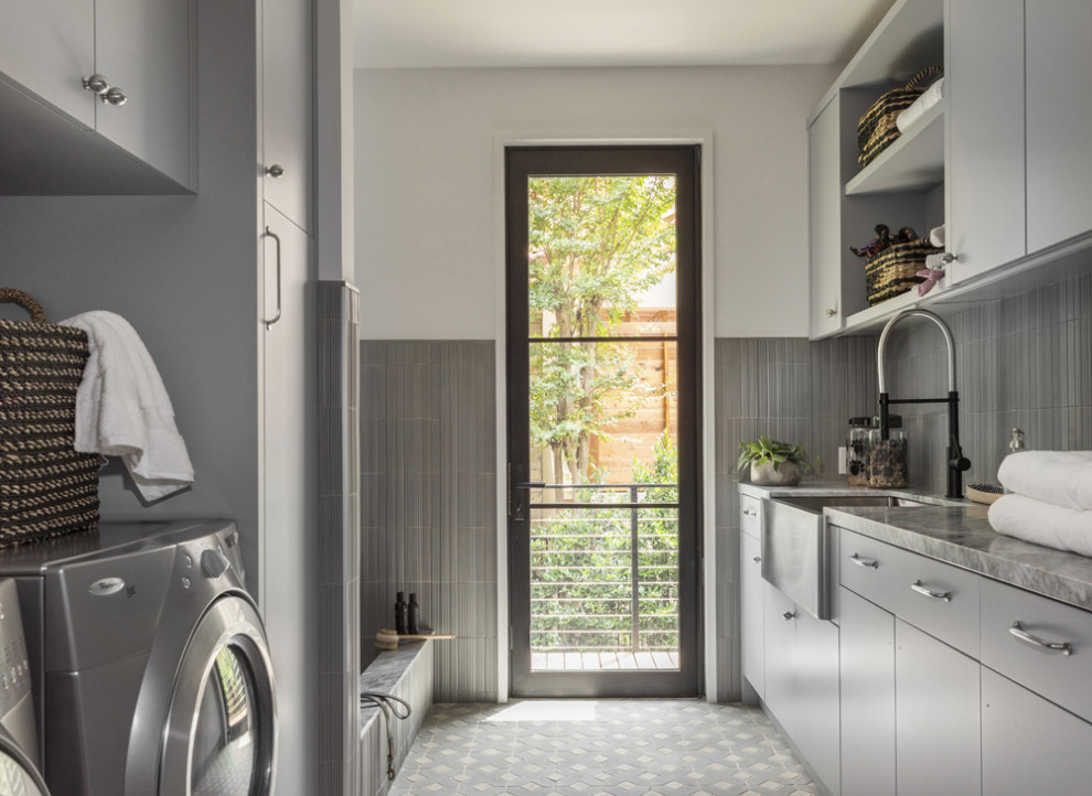 Inspiration for a mid-sized transitional galley utility room in Austin with a farmhouse sink, flat-panel cabinets, grey cabinets, marble benchtops, grey splashback, ceramic splashback, white walls, ceramic floors, a side-by-side washer and dryer, grey floor, grey benchtop and decorative wall panelling.
