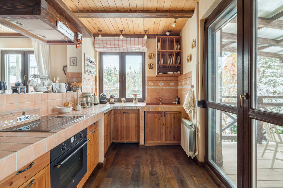 Photo of a country l-shaped eat-in kitchen in Moscow with a drop-in sink, recessed-panel cabinets, medium wood cabinets, tile benchtops, beige splashback, ceramic splashback, black appliances, dark hardwood floors, brown floor and a peninsula.