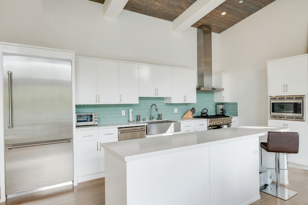 Inspiration for a beach style l-shaped kitchen in Los Angeles with a farmhouse sink, flat-panel cabinets, white cabinets, quartz benchtops, green splashback, matchstick tile splashback, stainless steel appliances, light hardwood floors, with island and beige floor.