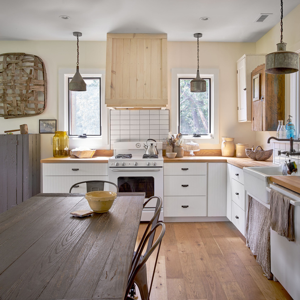 Design ideas for a mid-sized country l-shaped eat-in kitchen in Other with a farmhouse sink, white cabinets, wood benchtops, white appliances, no island, white splashback, medium hardwood floors, brown floor and brown benchtop.