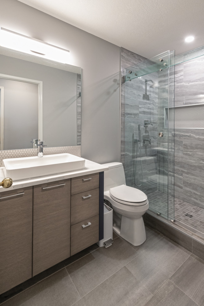 Design ideas for a medium sized modern ensuite bathroom in Edmonton with flat-panel cabinets, light wood cabinets, a freestanding bath, an alcove shower, white tiles, ceramic tiles, granite worktops, white worktops, a single sink, a built in vanity unit, a one-piece toilet, grey walls, ceramic flooring, a vessel sink, grey floors, a sliding door and panelled walls.