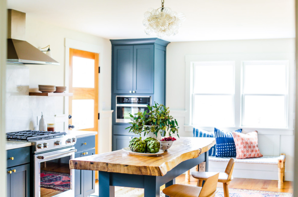 This is an example of a mid-sized eclectic l-shaped kitchen in Portland Maine with shaker cabinets, blue cabinets, granite benchtops, white splashback, stone slab splashback, stainless steel appliances, light hardwood floors, with island and white benchtop.