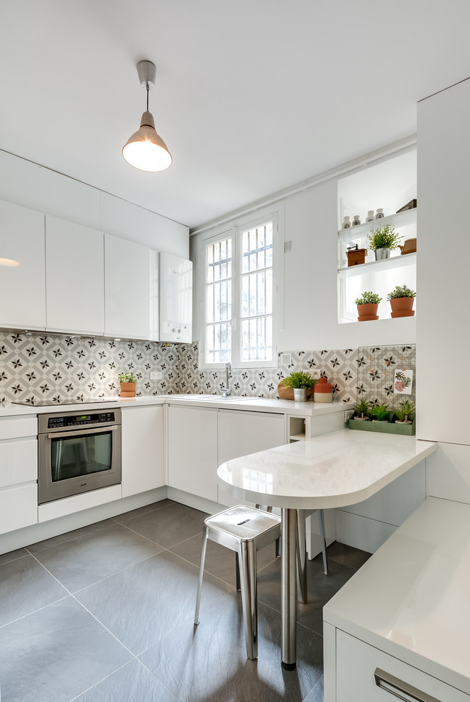 Design ideas for a mid-sized contemporary l-shaped eat-in kitchen in Montpellier with white cabinets, laminate benchtops, cement tile splashback, porcelain floors, with island, grey floor, white benchtop, a drop-in sink, flat-panel cabinets, multi-coloured splashback and stainless steel appliances.