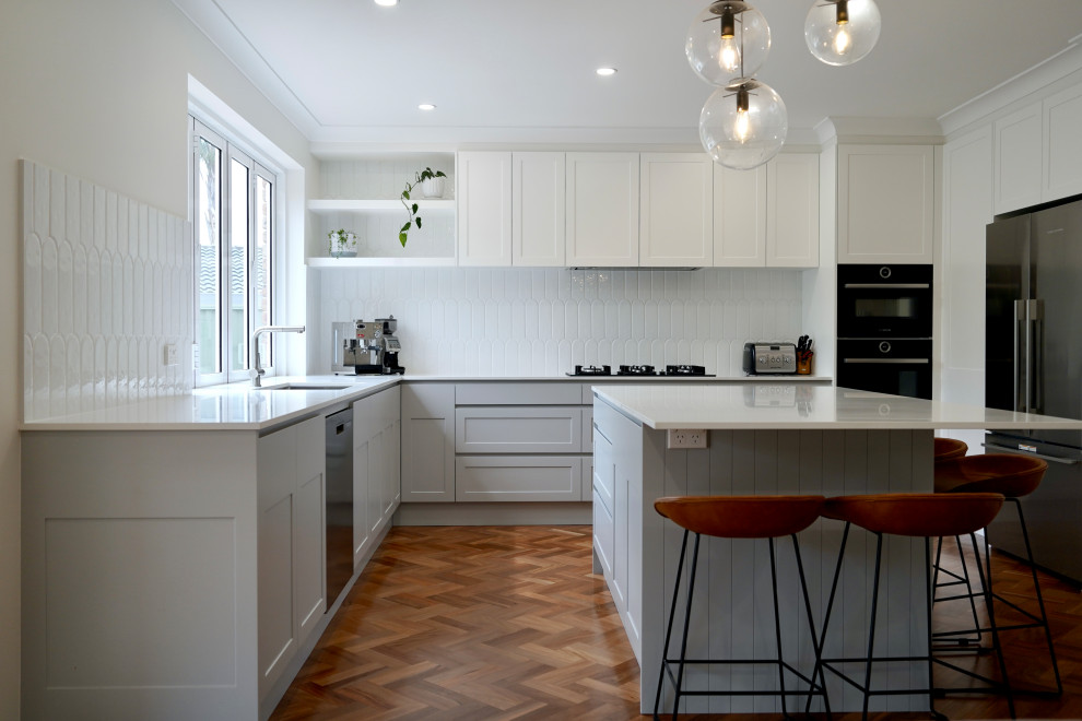 This is an example of a mid-sized transitional eat-in kitchen in Sydney with an undermount sink, shaker cabinets, grey cabinets, quartz benchtops, white splashback, ceramic splashback, black appliances, medium hardwood floors, with island, brown floor and white benchtop.