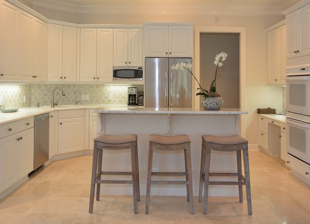 This is an example of a beach style u-shaped eat-in kitchen in Miami with white cabinets, beige splashback, white appliances, with island, an undermount sink, travertine floors and stone tile splashback.
