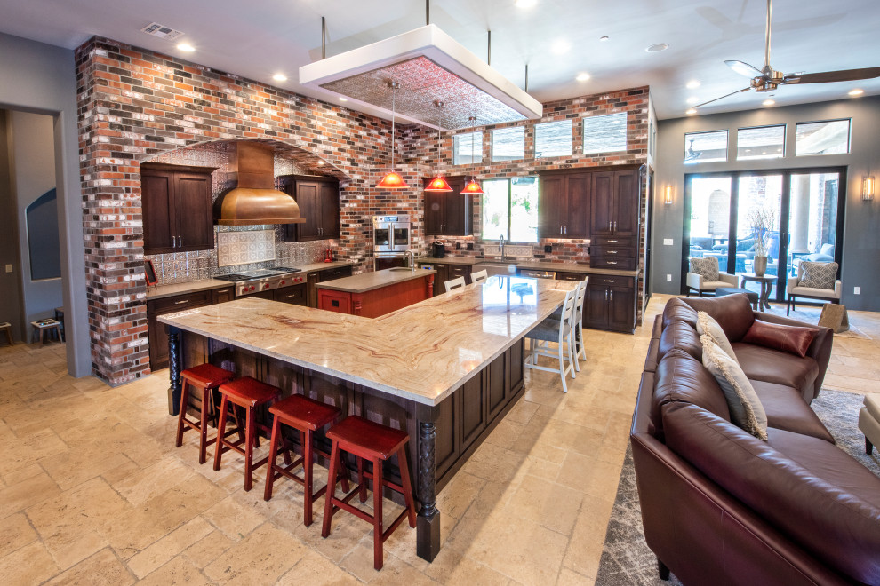 This is an example of an expansive arts and crafts l-shaped open plan kitchen in Phoenix with a farmhouse sink, shaker cabinets, brown cabinets, granite benchtops, metallic splashback, metal splashback, stainless steel appliances, travertine floors, multiple islands, beige floor and red benchtop.