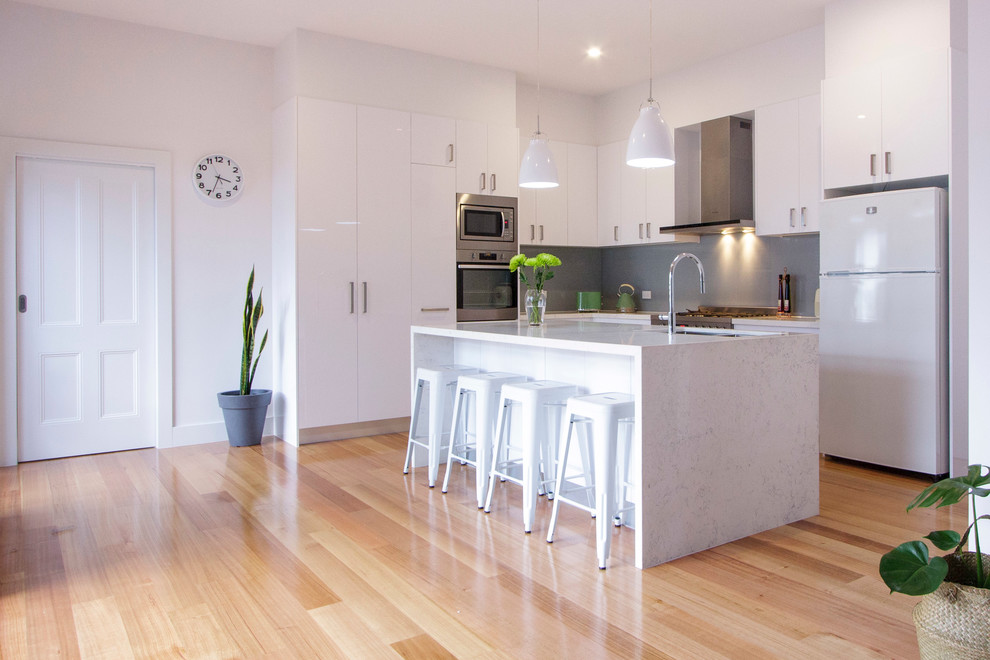 Inspiration for a contemporary l-shaped open plan kitchen in Melbourne with an undermount sink, white cabinets, quartz benchtops, grey splashback, glass sheet splashback, stainless steel appliances, light hardwood floors and with island.
