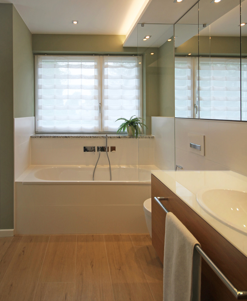 Duschbad With flächenbündigen Schrankfronten, hellbraunen Holzschränken, Einbaubadewanne, bodengleicher Dusche, Wandtoilette, weißen Fliesen, Keramikfliesen, grüner Wandfarbe, Einbauwaschbecken, Glaswaschbecken/Glaswaschtisch, braunem Boden and Falttür-Duschabtrennung in Munich